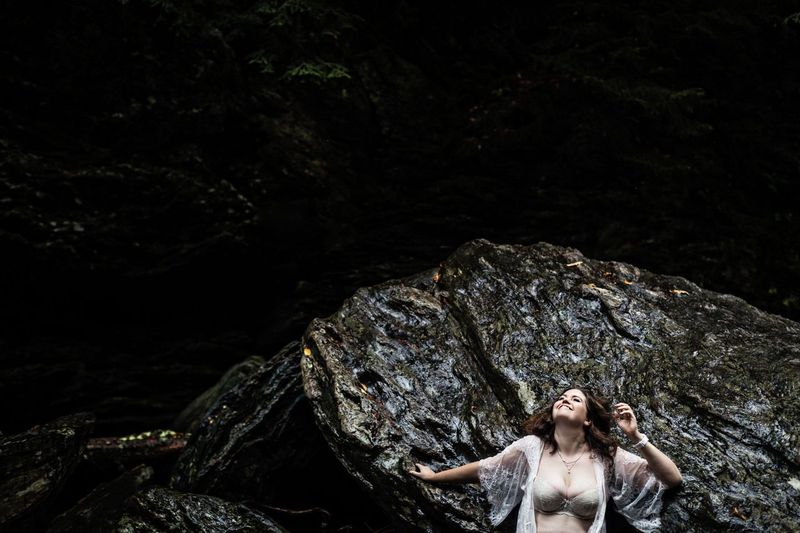 A woman in a sheer white garment leans against a large, textured rock formation. She gazes upward with a serene expression, surrounded by a dark, forested background.