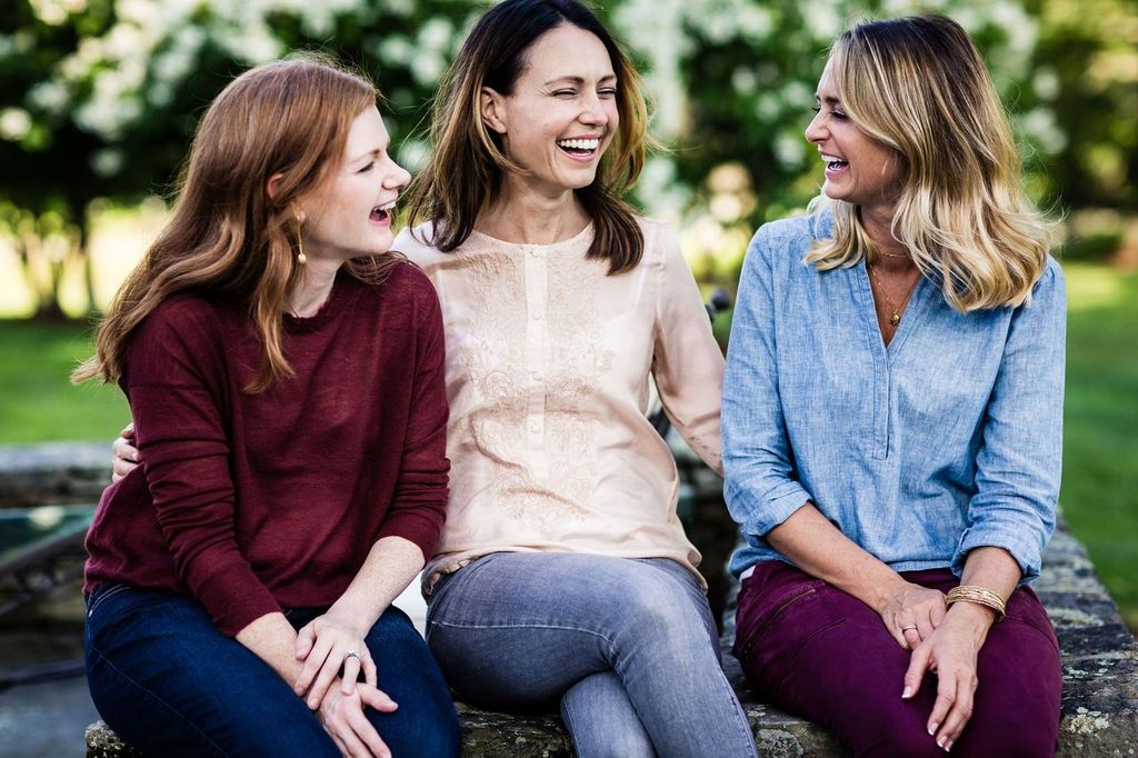Three women are sitting and laughing together outdoors. They are casually dressed, with two wearing jeans and one in maroon pants. The background features blurred greenery, suggesting a park or garden setting.