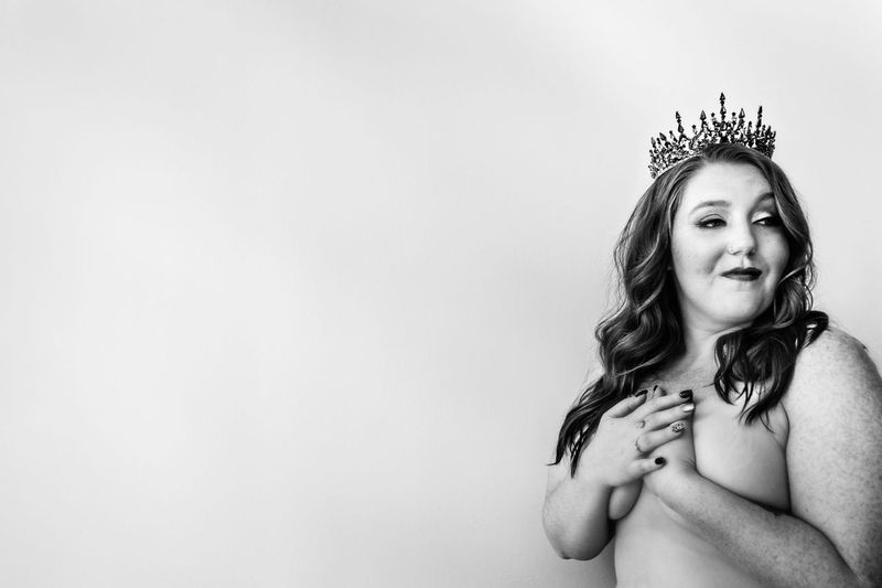 A woman wearing a tiara poses confidently with a slight smile, covering her chest with her hands, against a plain backdrop. The image is in black and white, highlighting her expression and the intricate design of the tiara.