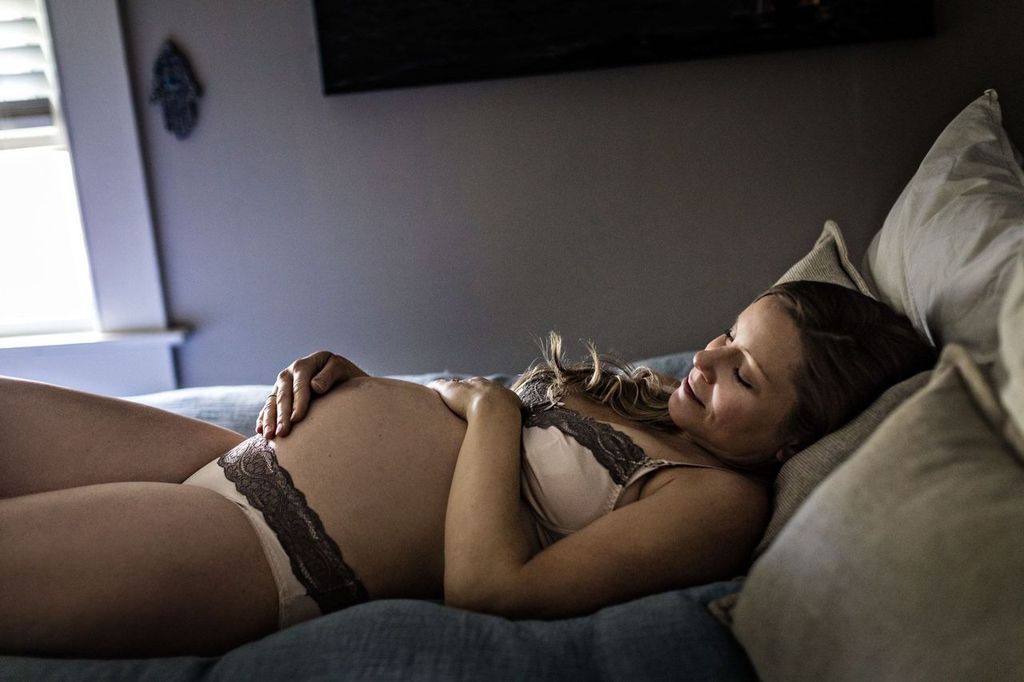 A pregnant woman is lying on a bed, resting her hands on her belly. She appears relaxed and peaceful, wearing underwear with lace details. The room is softly lit, creating a serene atmosphere.