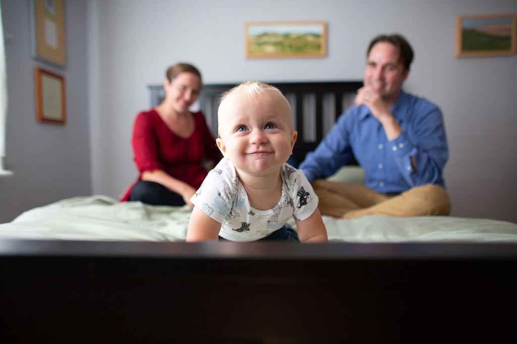 A smiling toddler in a patterned shirt crawls on a bed, while a man and woman sit behind, blurred in the background. The room has soft lighting, with framed pictures on the walls.
