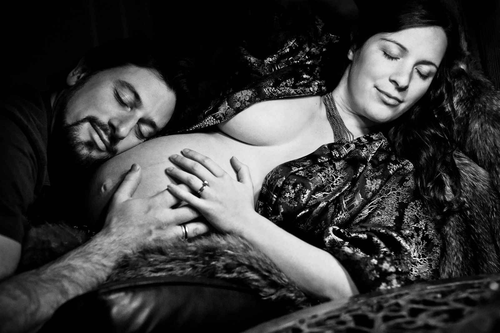 A black and white photo of a couple. A pregnant woman lies on her back with eyes closed, wearing a patterned robe. A man rests his head on her belly, smiling, with his hand gently placed on it. The scene conveys warmth and tenderness.