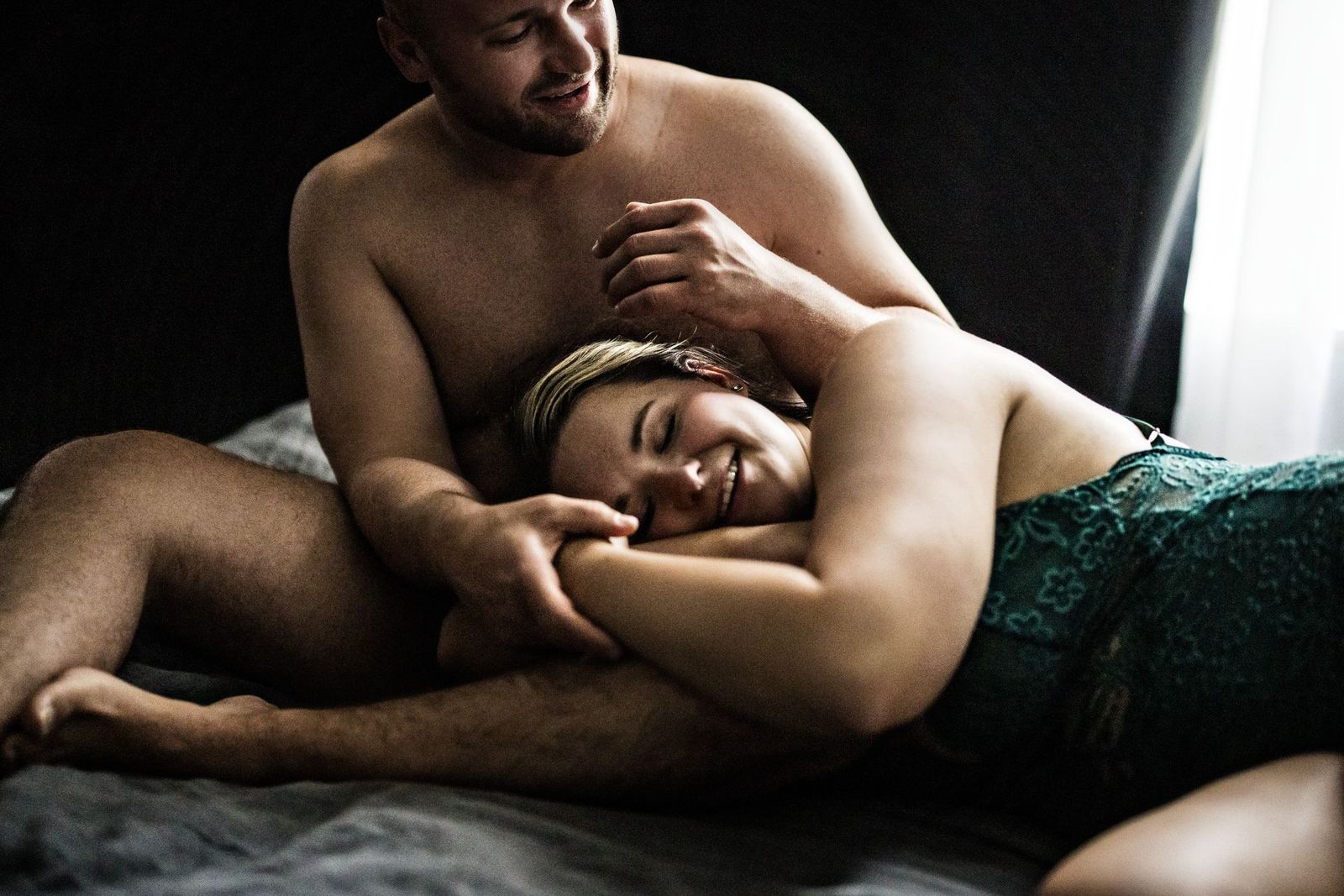 A couple is lying in bed together. The man is shirtless, smiling, and gently holding the womans arm. The woman is wearing a green top and resting her head on his leg, smiling peacefully. The setting is intimate and softly lit.