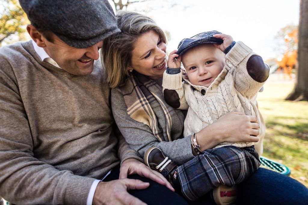 A happy family sits on a bench outdoors. The mother and father, both wearing sweaters and hats, smile at their young child, who is also wearing a cap and plaid pants. The toddler smiles and touches their cap. The background shows trees on a sunny day.