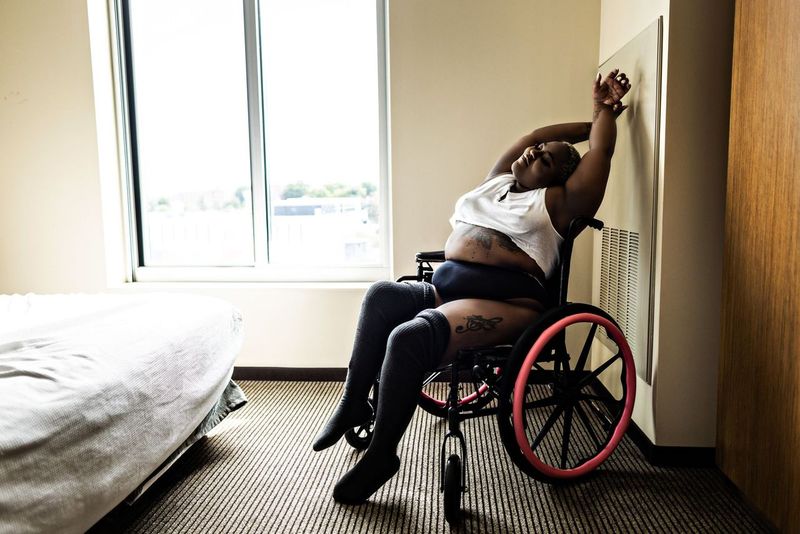 Woman in a wheelchair stretches with arms above their head in a room with a large window and a bed. They wear a sleeveless top, shorts, and knee-high socks. A tattoo is visible on their abdomen.