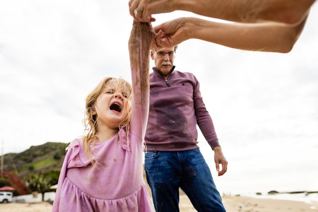 A young girl in a pink dress looks up excitedly at an adult holding a large fish. An older man stands in the background on a sandy beach with hills and cloudy sky visible.