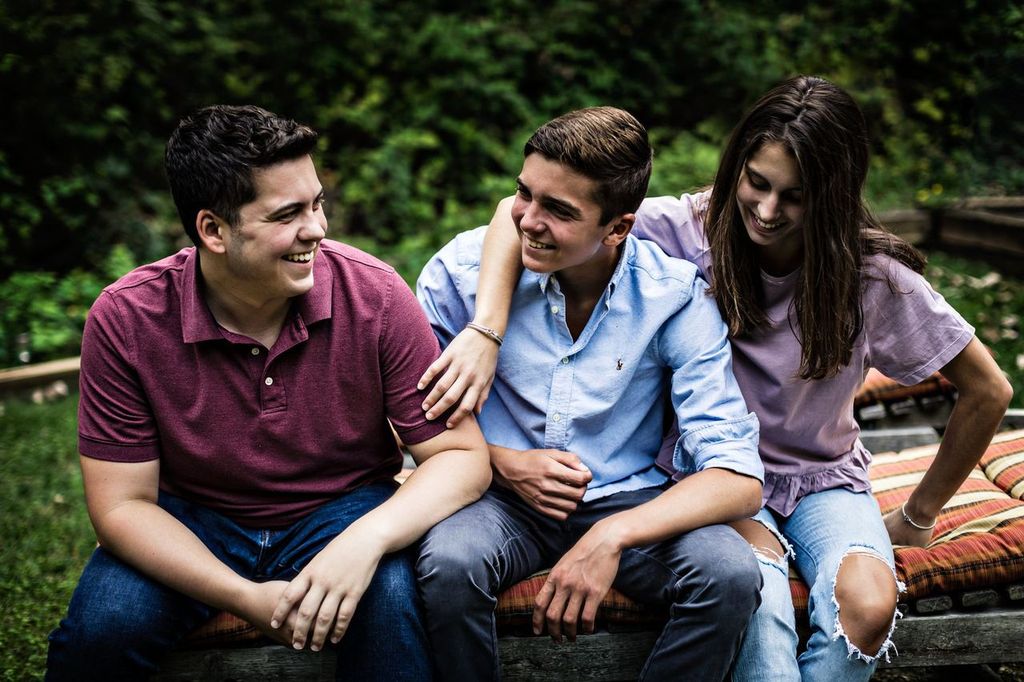 Three young adults sit outdoors on a bench, smiling and talking. The person on the left wears a red polo, the middle person is in a blue shirt, and the person on the right wears a purple top and ripped jeans. Green foliage is in the background.