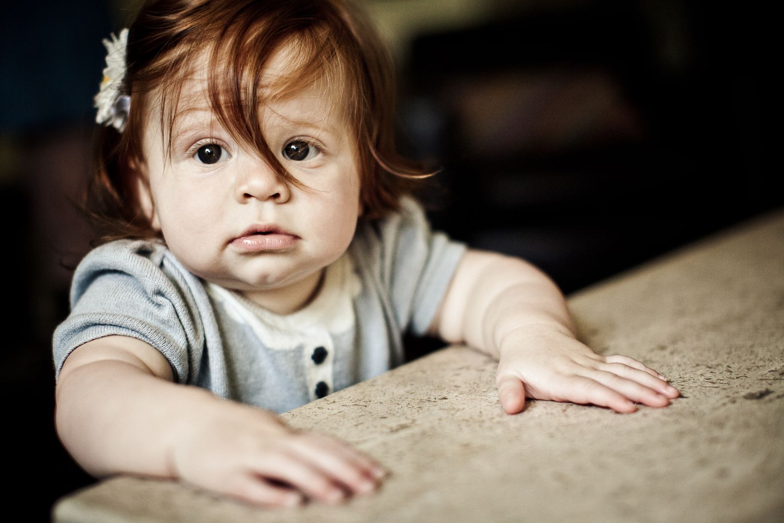 A toddler with red hair and a flower hairpin leans on a table, wearing a gray outfit with a white collar. Her wide eyes look slightly to the side, and her hands rest on the table surface. The background is softly blurred.