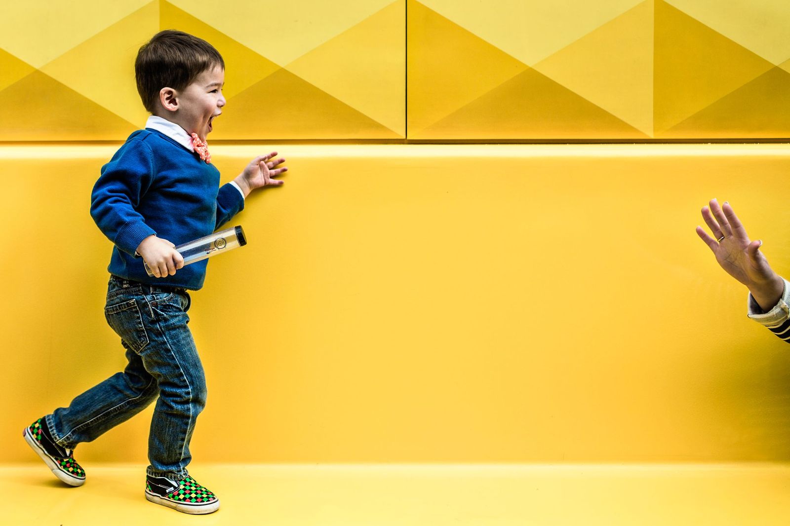 A smiling child in a blue sweater and jeans runs across a bright yellow background, holding a small tube. A persons hand reaches out towards the child from the right side of the image.