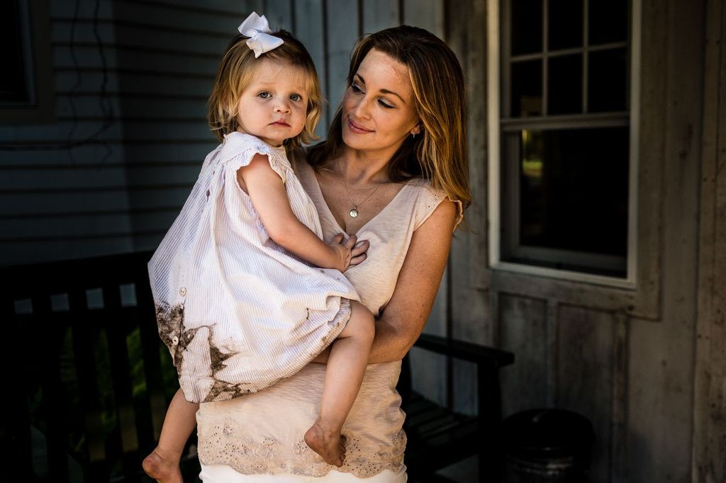 A woman holds a young girl dressed in a light, slightly dirt-stained dress with a bow in her hair. They stand near a wooden building with a bench and window in the background. The woman wears a sleeveless top and looks at the child with a gentle expression.