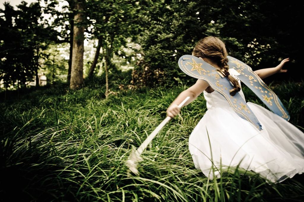 A child wearing fairy wings and a white dress is running through a lush, grassy area in a forest. They are holding a wand, and their hair is tied back. The scene is whimsical and filled with greenery.