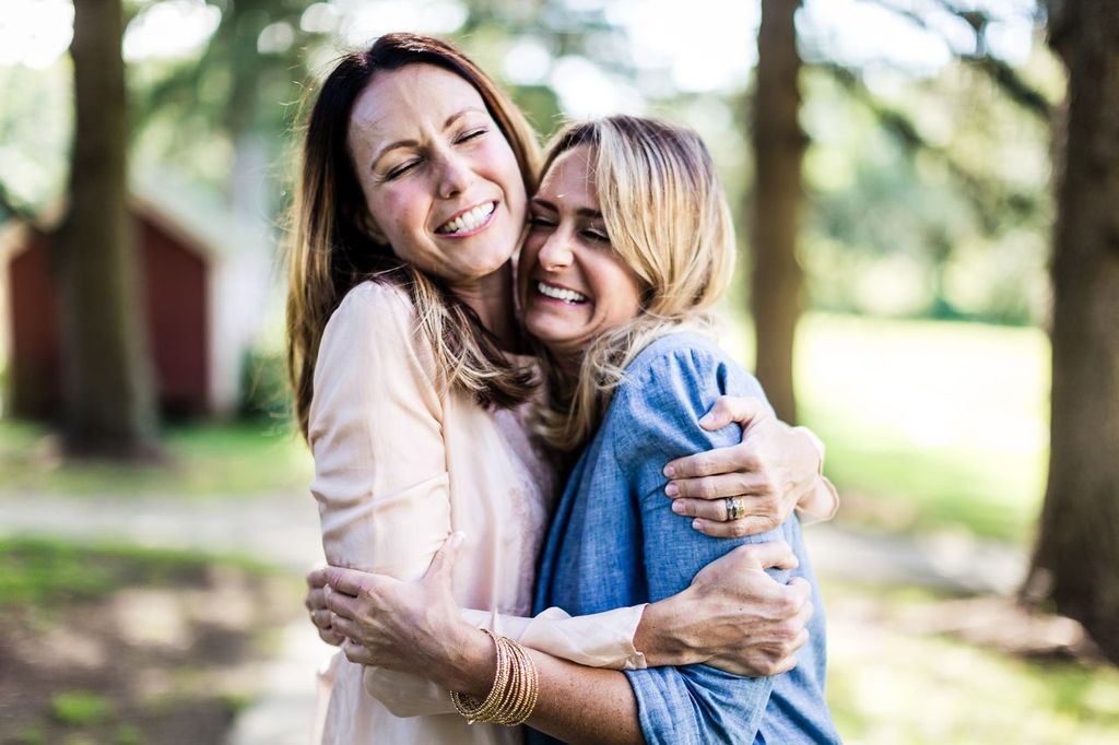 Two women are embracing and smiling joyfully in an outdoor setting with trees and blurred background. One is wearing a light pink top and the other a blue shirt. Their expressions convey happiness and friendship.