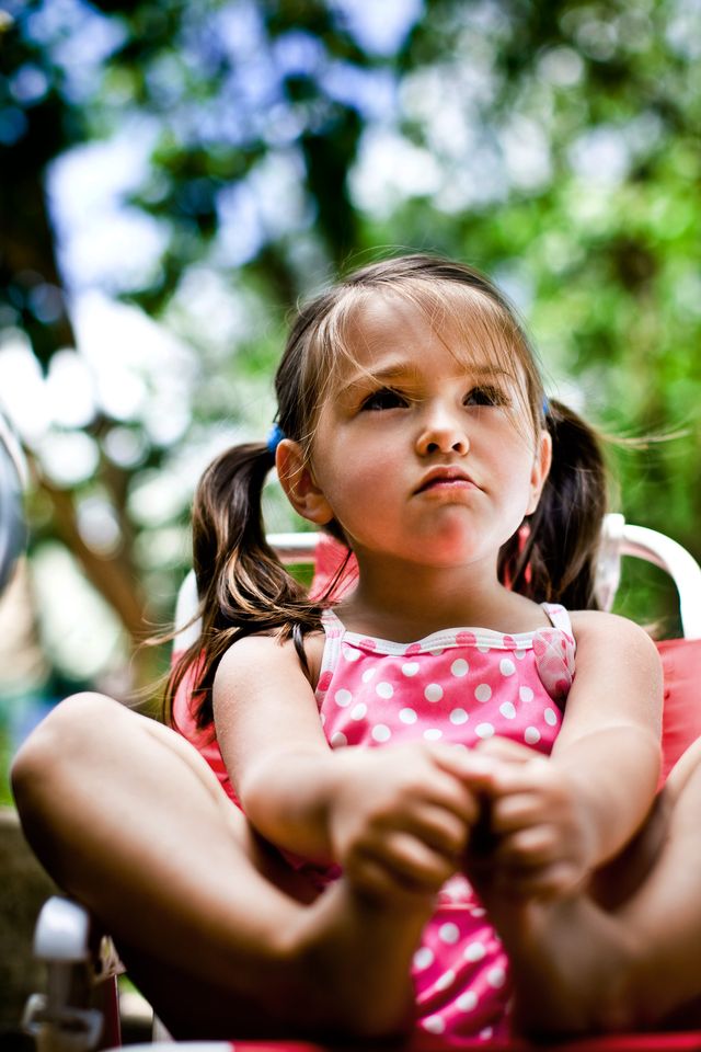 A young girl with pigtails sits on a chair outdoors, wearing a pink polka-dot outfit. She has a contemplative expression, with lush greenery blurred in the background.