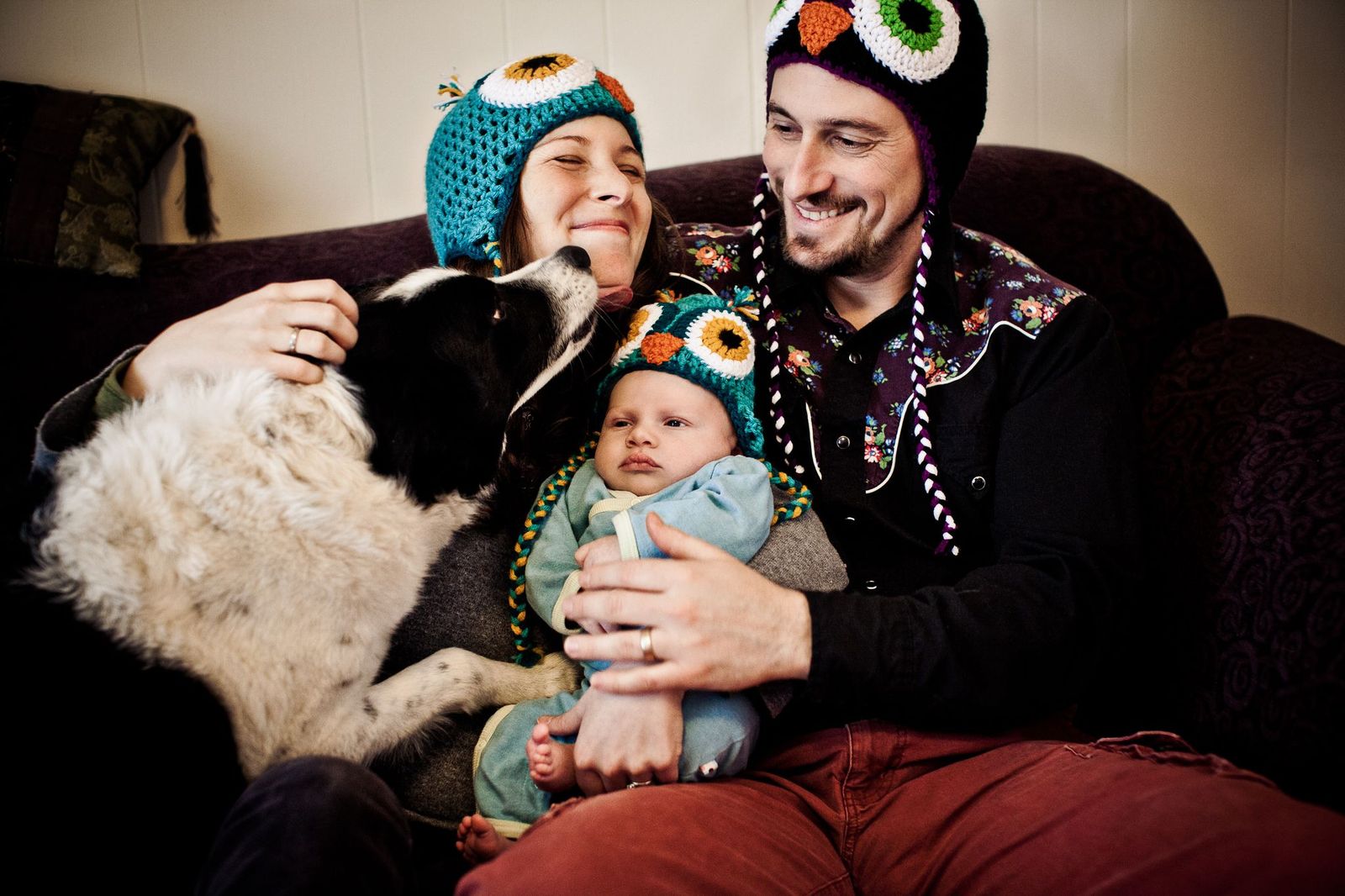 A family of three, wearing colorful owl hats, sits on a couch. The woman holds a baby dressed in blue, while the man embraces them both. A black and white dog gently reaches towards the womans face, adding to the warmth of the scene.