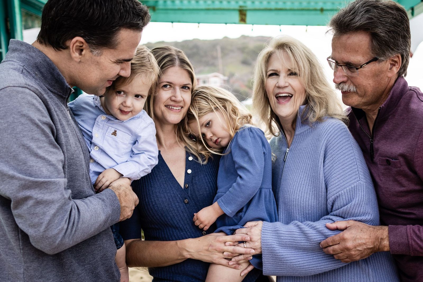 A group of six people, including two adults and two children, pose closely together under a green structure. The adults are smiling, and the children are being held. They are in winter outfits, and the background shows a hilly landscape.