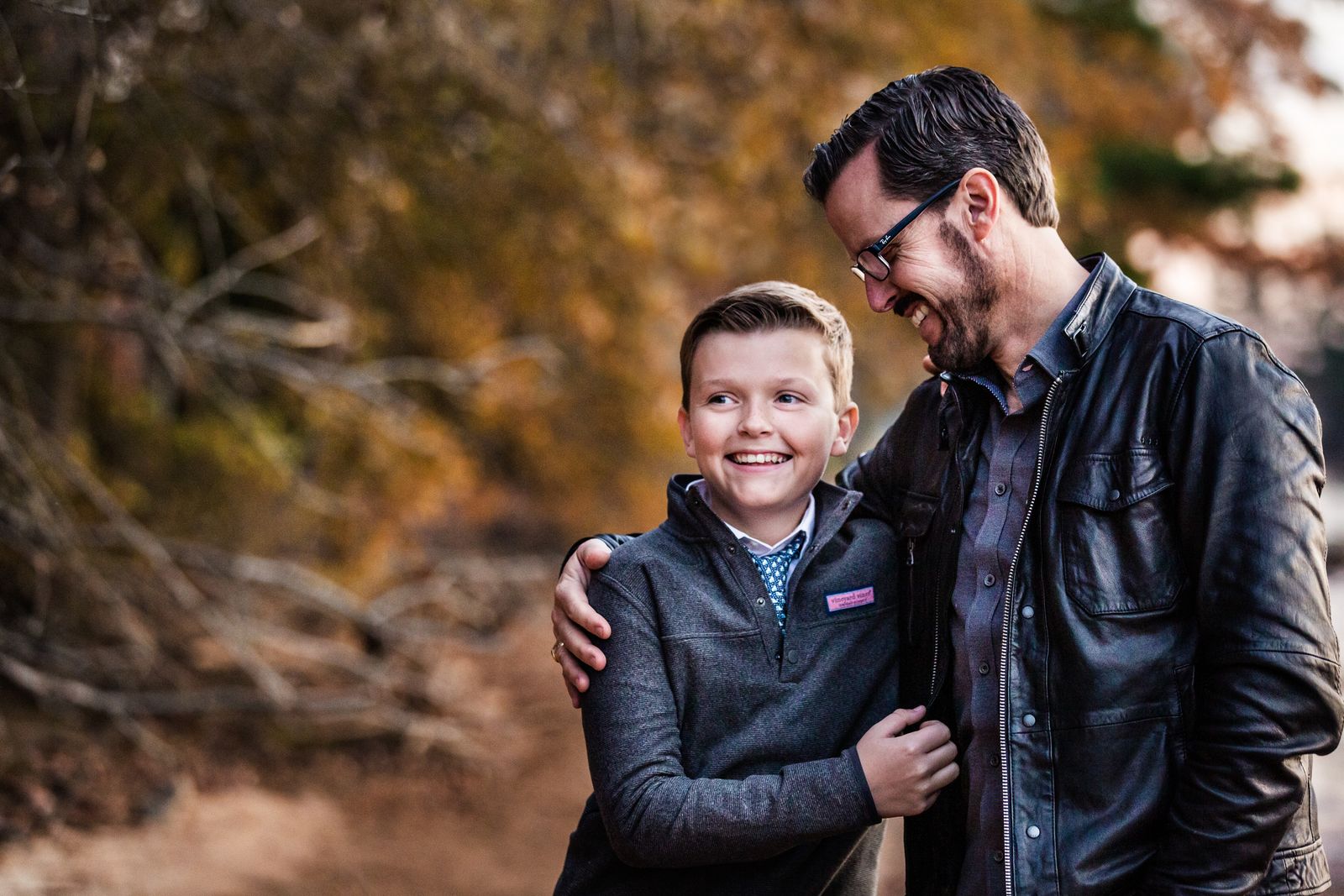 A man in a leather jacket and glasses affectionately embraces a smiling boy wearing a gray sweater. They stand outdoors with autumn foliage in the background, sharing a joyful moment together.