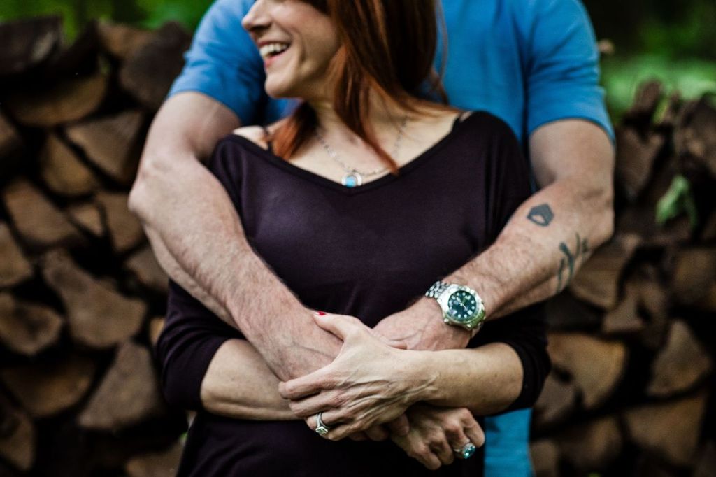 A person with long red hair is being embraced from behind by someone wearing a blue shirt, displaying tattoos and a watch. They stand in front of a stack of firewood, both appearing joyful.