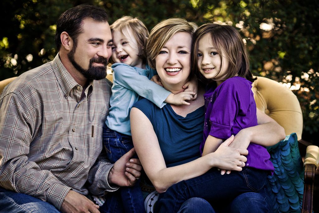 A family of four sitting together on a sofa outdoors. The father has a beard and wears a plaid shirt. The mother has short hair and wears a blue top. They are smiling with their two young children, a boy in blue and a girl in purple.