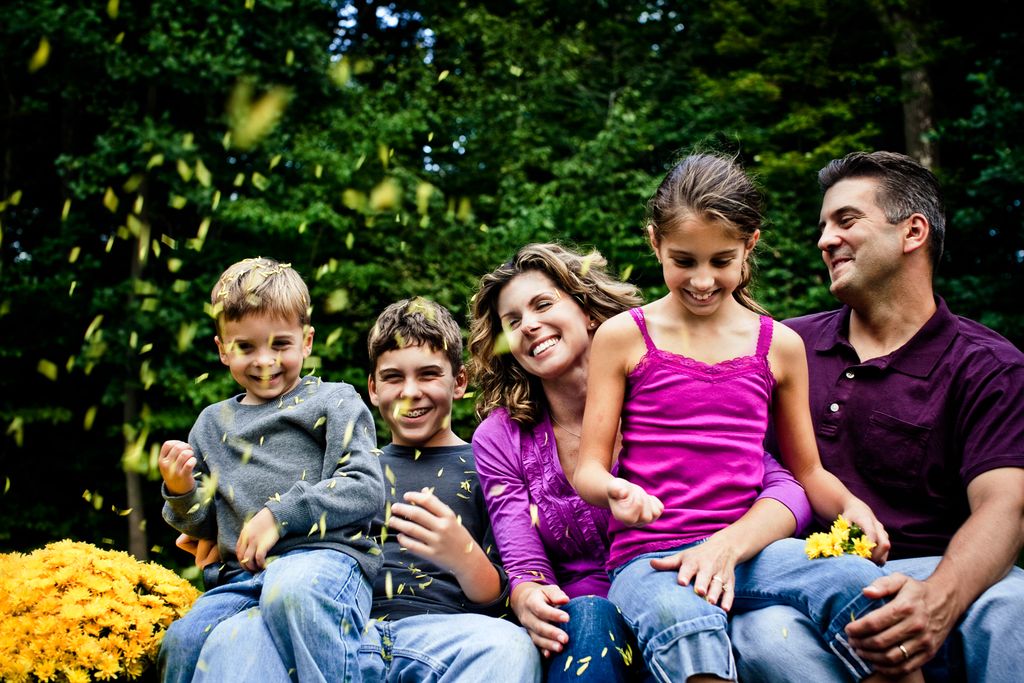 A family of five, including two boys, a girl, and two adults, sits together outside surrounded by greenery. They are smiling and playing with yellow flower petals, creating a joyful and relaxed atmosphere.