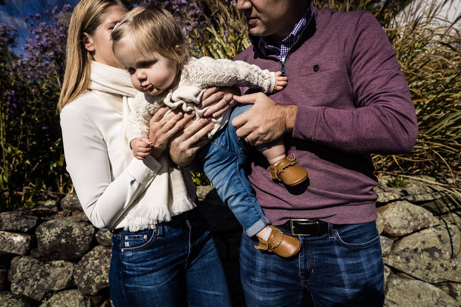 A toddler wearing a knit sweater and jeans is held by two adults. The adult on the left is holding the childs arm, while the adult on the right supports the childs leg. They are standing outdoors with foliage and rocks in the background.