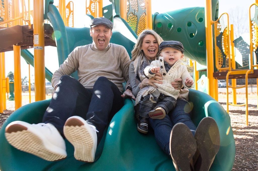 A family of three enjoys a ride on a green playground slide. The mother, father, and child are smiling and wearing winter clothing and hats. The father and son wear matching caps. Bright playground equipment is visible in the background.