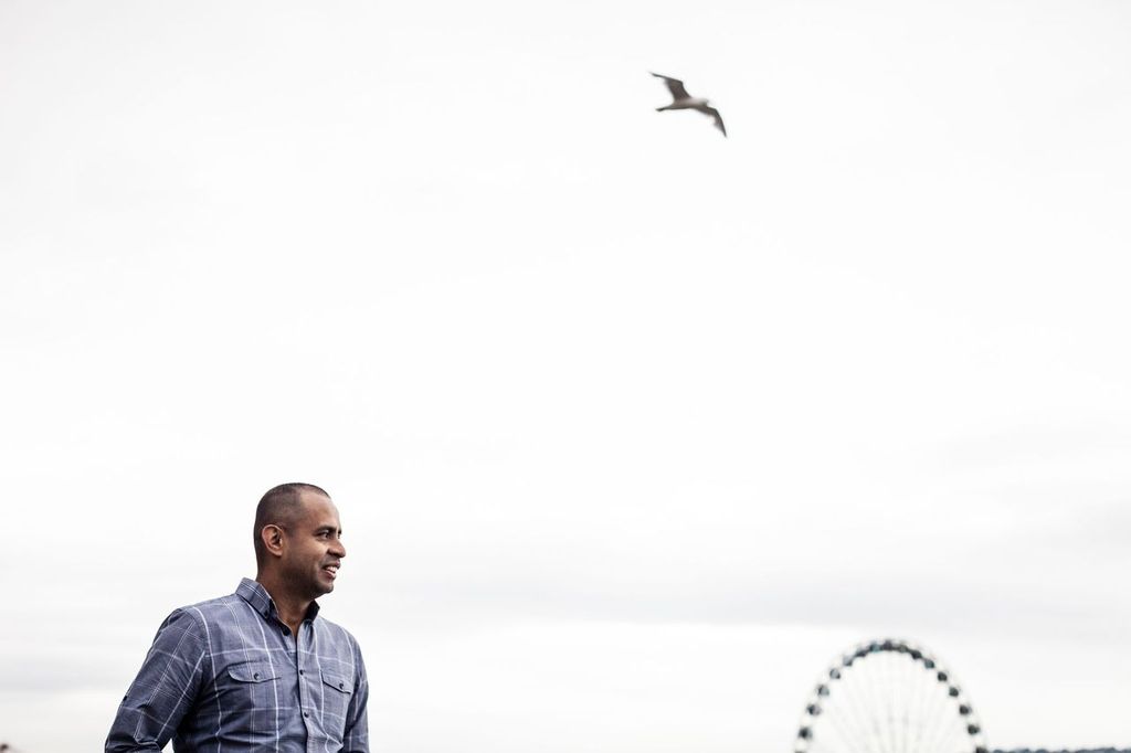 A person in a blue plaid shirt stands outdoors with a Ferris wheel in the background. A seagull flies above in the cloudy sky.