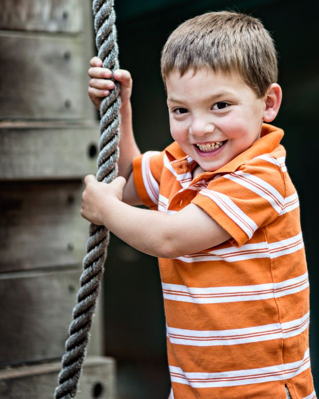 A young boy wearing an orange and white striped shirt smiles while holding onto a rope. He is standing near a wooden structure, possibly part of a playground. The background is blurred, focusing attention on the boys joyful expression.