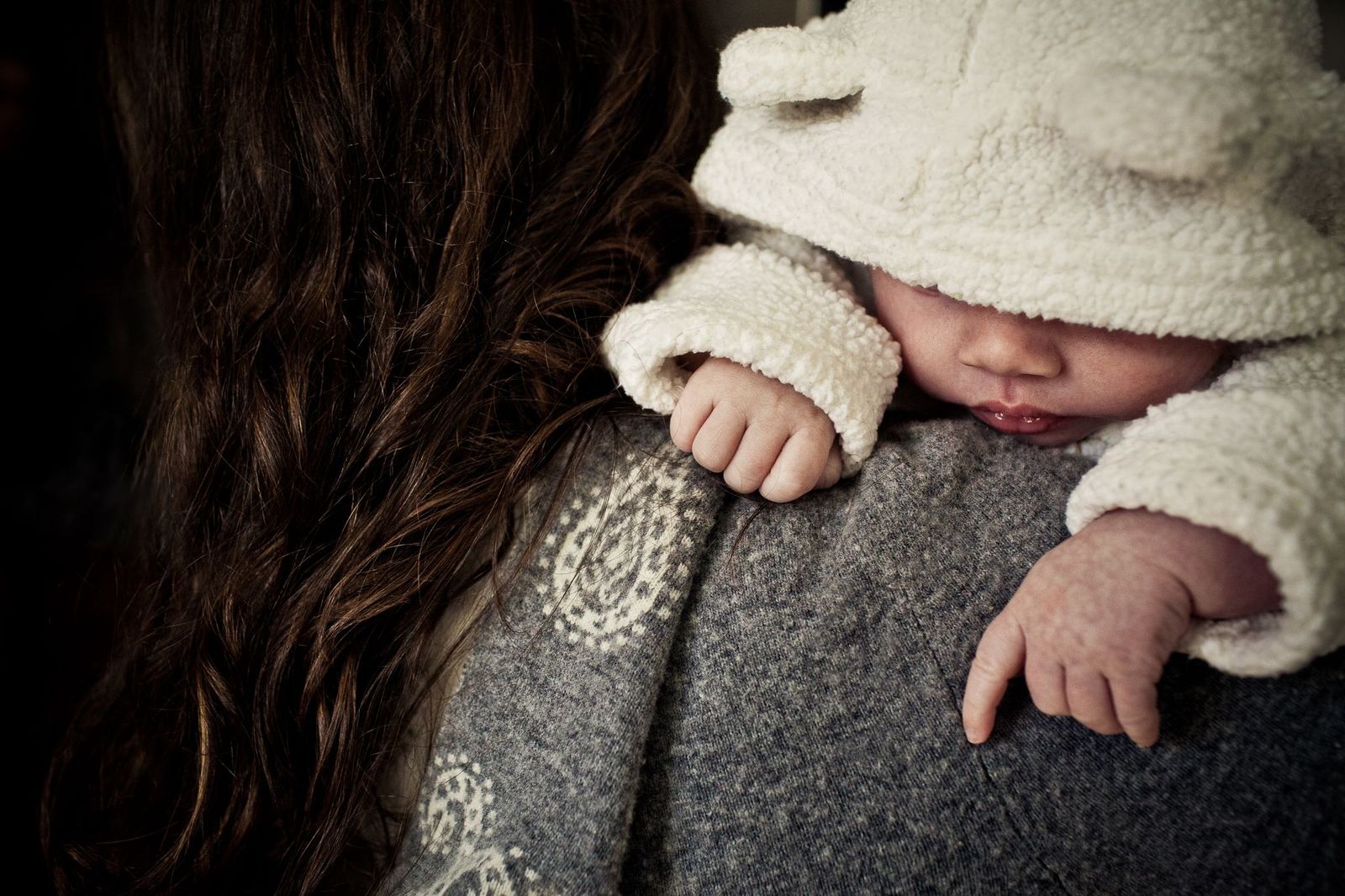 A baby wearing a fluffy hooded outfit rests their head on a persons shoulder. The baby has chubby cheeks and their hands are visible, lightly gripping the persons patterned sweater. The persons long brown hair is seen from the back.
