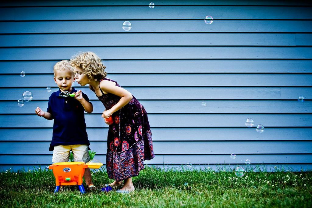 Two children playing outside in the grass with bubbles. One child, wearing a dark blue shirt, holds a bubble wand, while the other, in a patterned dress, leans in to blow bubbles. A blue wall forms the background.