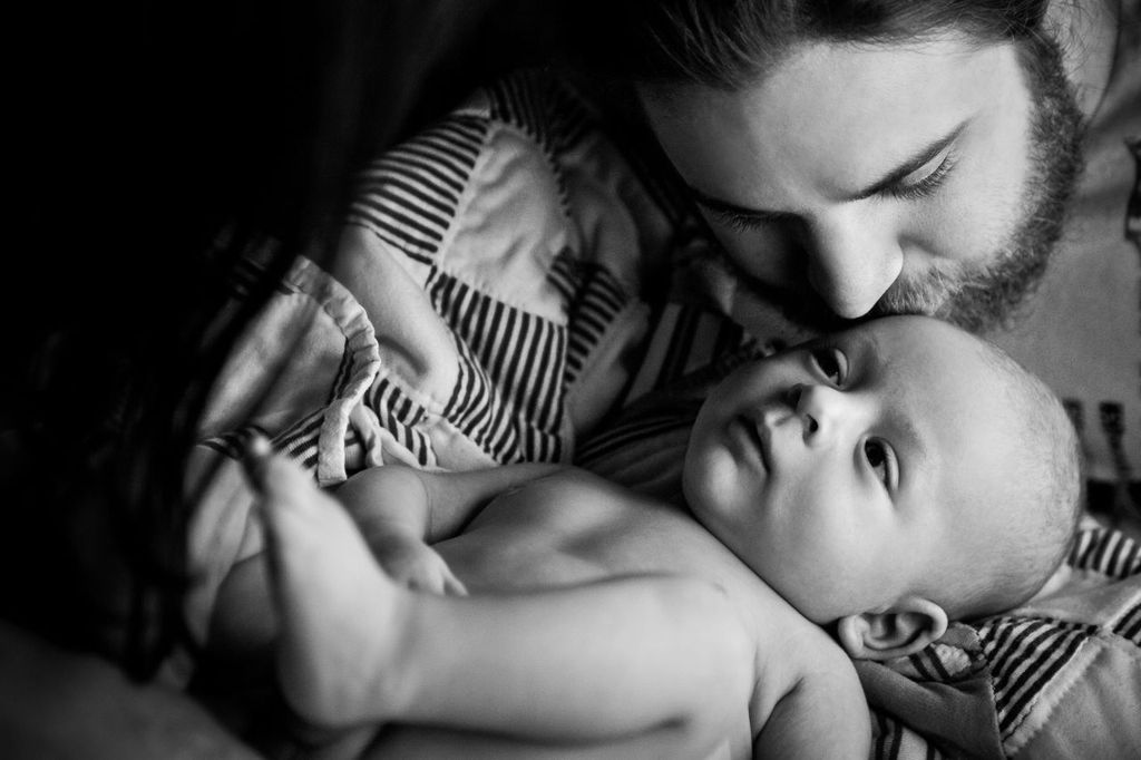 A black and white image of a person gently cuddling and looking at an infant who is lying on a patterned blanket. The adult has a beard, and the baby looks calm and nestled close.