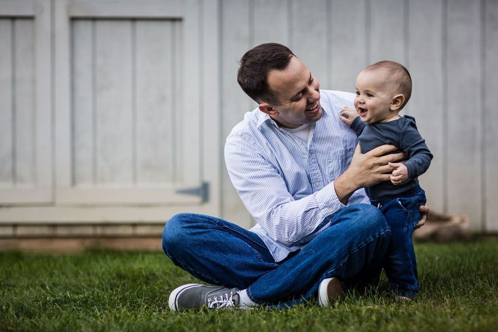 A man sits on the grass, wearing a light blue shirt and jeans, smiling at a standing baby who is dressed in a dark grey shirt and jeans. They are in front of a light-colored wooden fence.