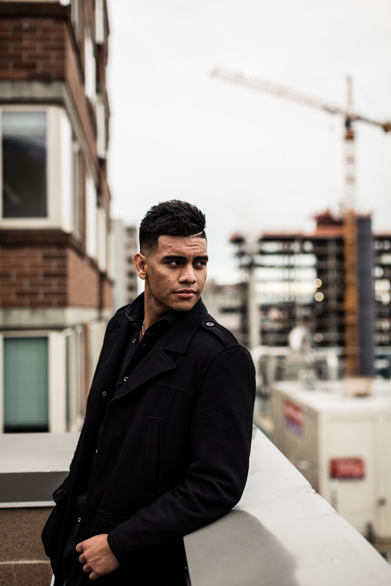 A person stands on a balcony, wearing a black coat and looking to the side. The background shows a partially constructed building with a crane, and surrounding structures under a cloudy sky.