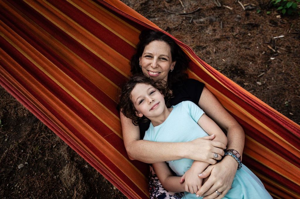 A woman and a child lying together in a colorful hammock, smiling at the camera. The woman has dark curly hair and is wearing a black shirt, while the child has light curly hair and is wearing a light blue shirt. Surrounding them is a forest floor.