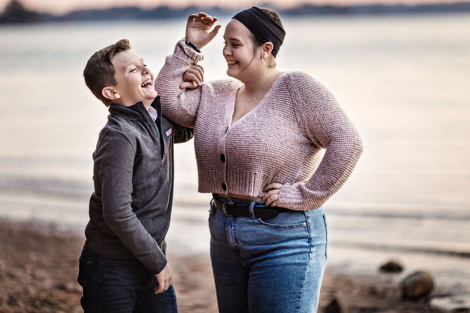 A boy and a young woman laugh together by the waters edge. The boy wears a grey jacket and jeans, while the woman is in a pink sweater and jeans with a black headband. The shoreline and water are blurred in the background.