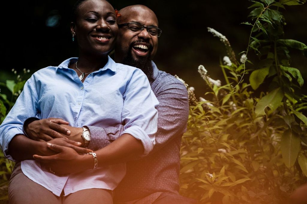 A couple sits closely together, smiling and wrapping their arms around each other. They are surrounded by green foliage and white flowers, creating a serene outdoor setting.