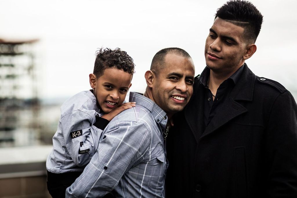 Three people stand together outdoors, smiling. A young child in a light gray shirt is held by a middle-aged man in a blue plaid shirt. Next to them is another man wearing a dark coat. The background is blurred, showing a cloudy sky and a building.
