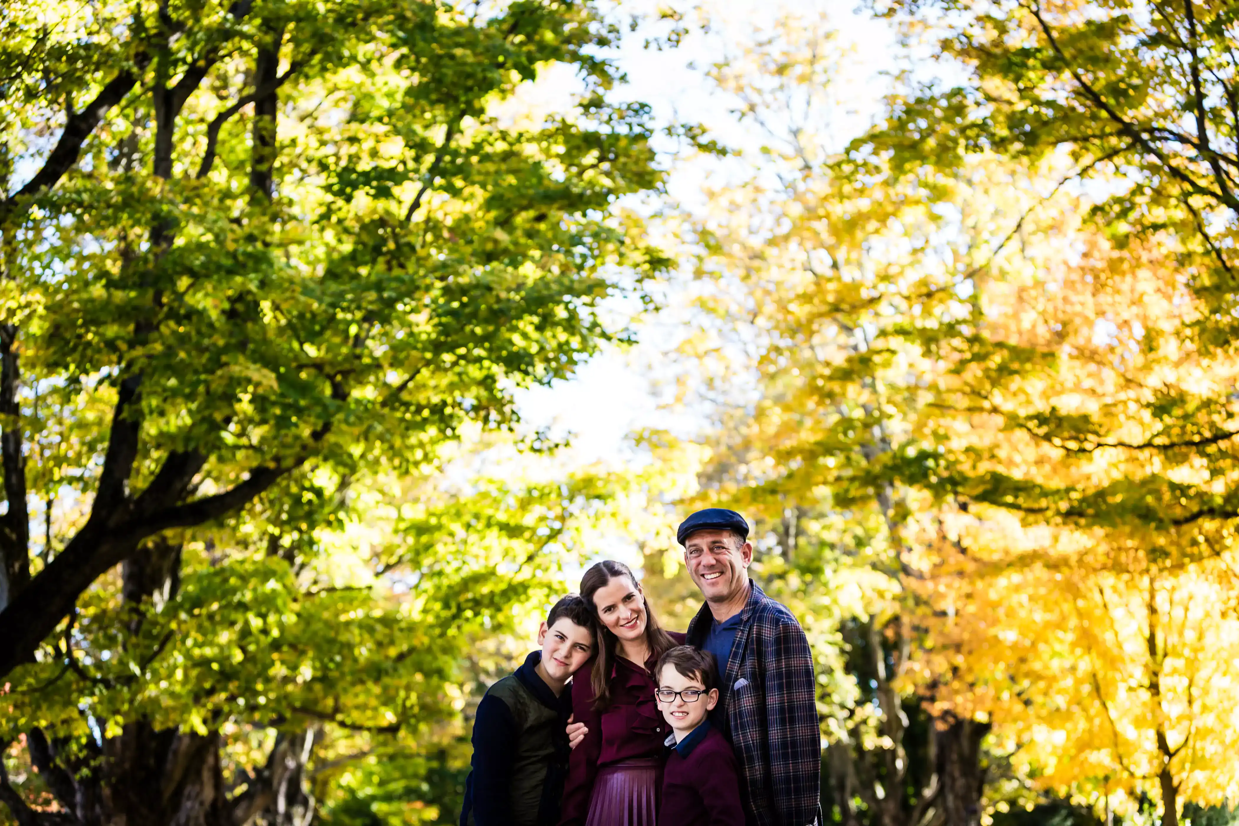 A family of four stands close together, smiling under a canopy of bright green and yellow autumn leaves. The sun filters through the trees, creating a warm and cheerful atmosphere.