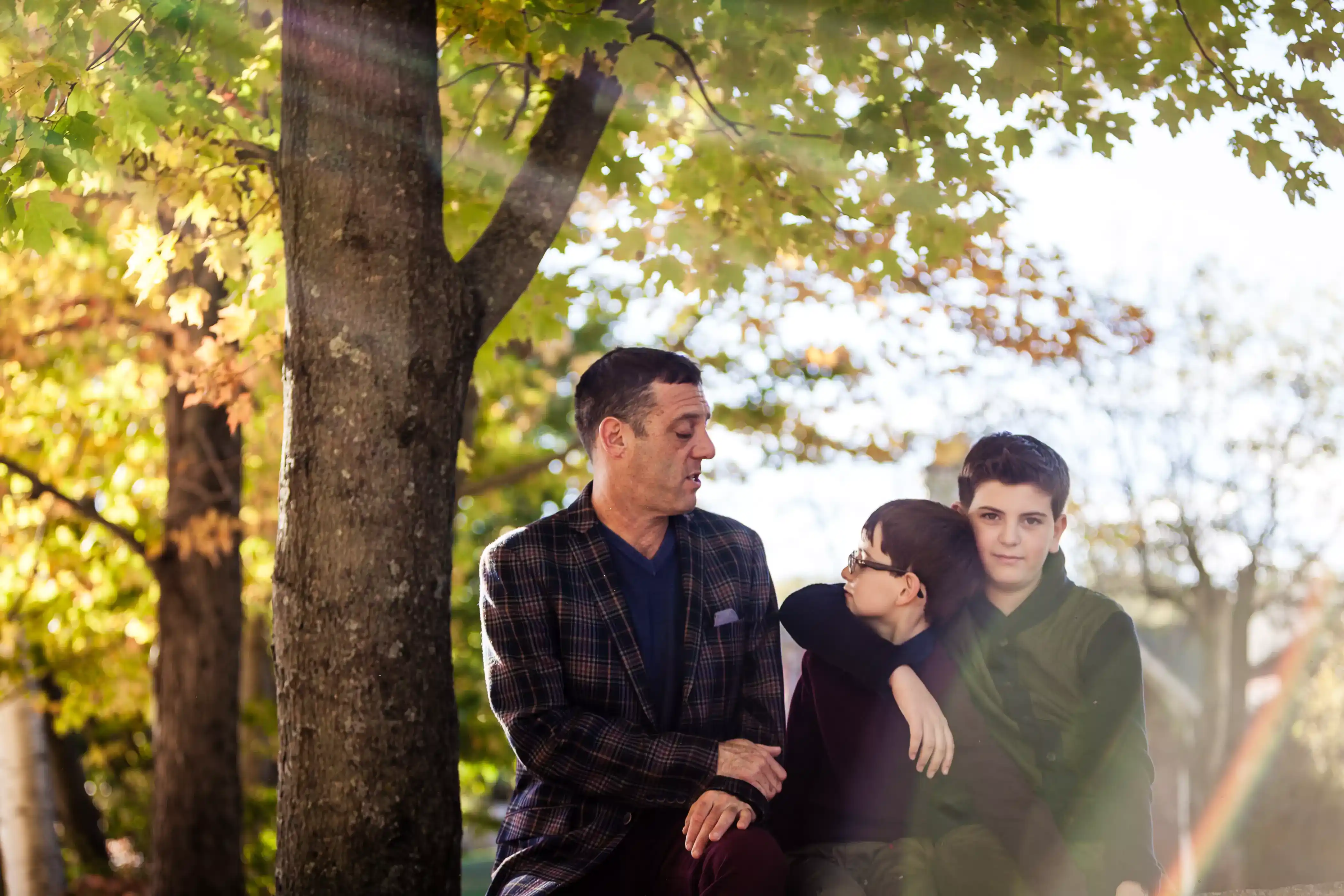 A man and two boys sit together on a log under a tree with autumn leaves. The man and the boy in the middle make eye contact and smile, while the other boy looks at the camera. Sunlight filters through the branches, casting a warm glow.