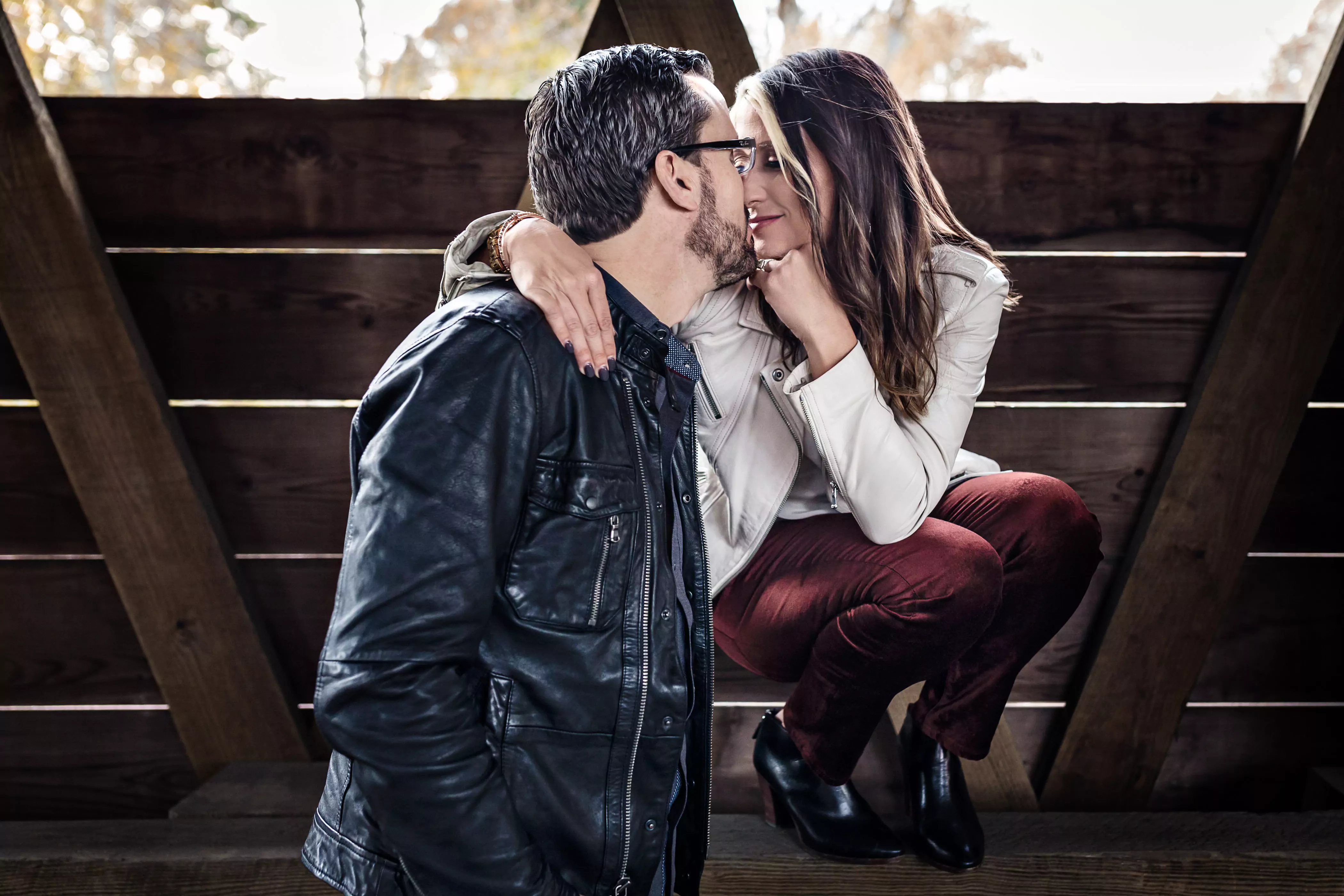 A couple is seated closely in an intimate pose. The man, in a black leather jacket and glasses, leans towards the woman, who is wearing a white jacket and maroon pants. She crouches playfully, touching his chin as they hold eye contact.