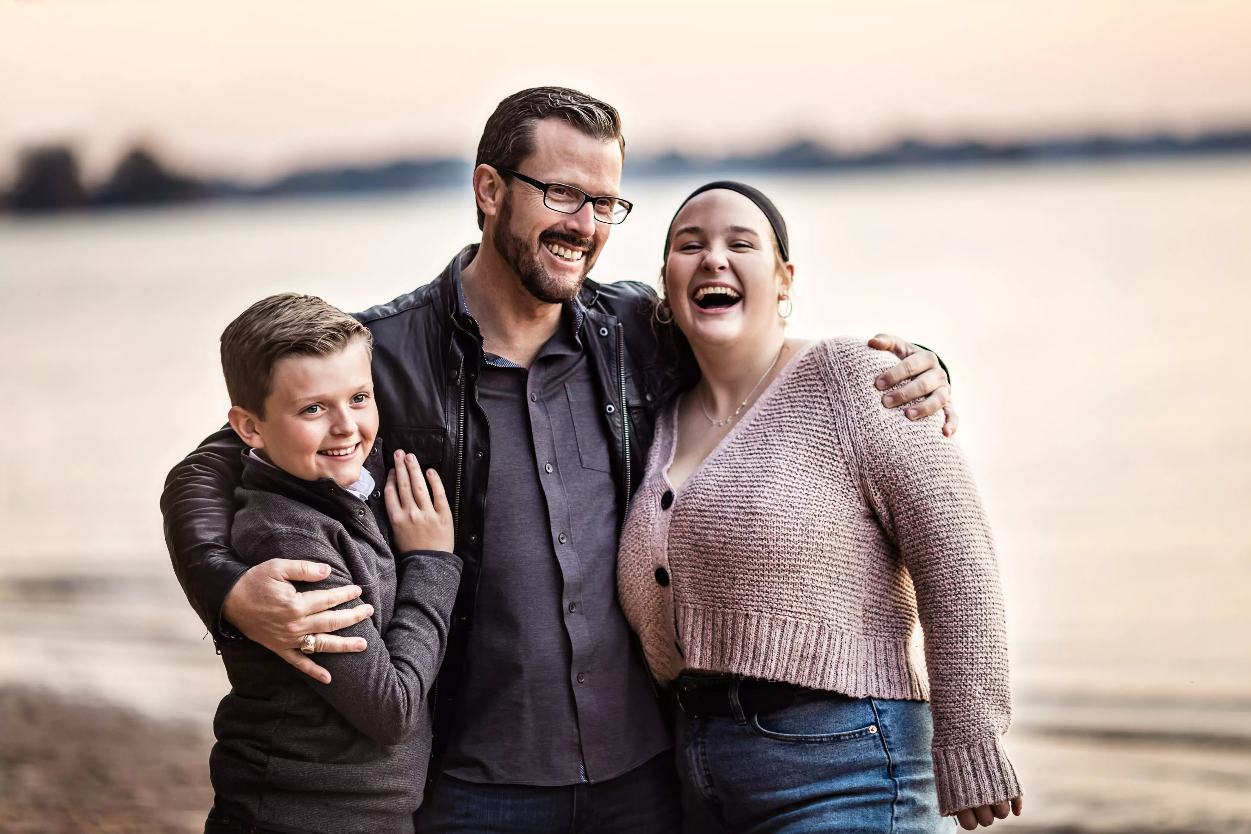 A man stands on a beach with a boy and a woman, all smiling joyfully. The man has a beard and glasses, wearing a dark jacket. The boy is in a gray sweater, and the woman wears a light sweater. The background features water and a blurred shoreline.