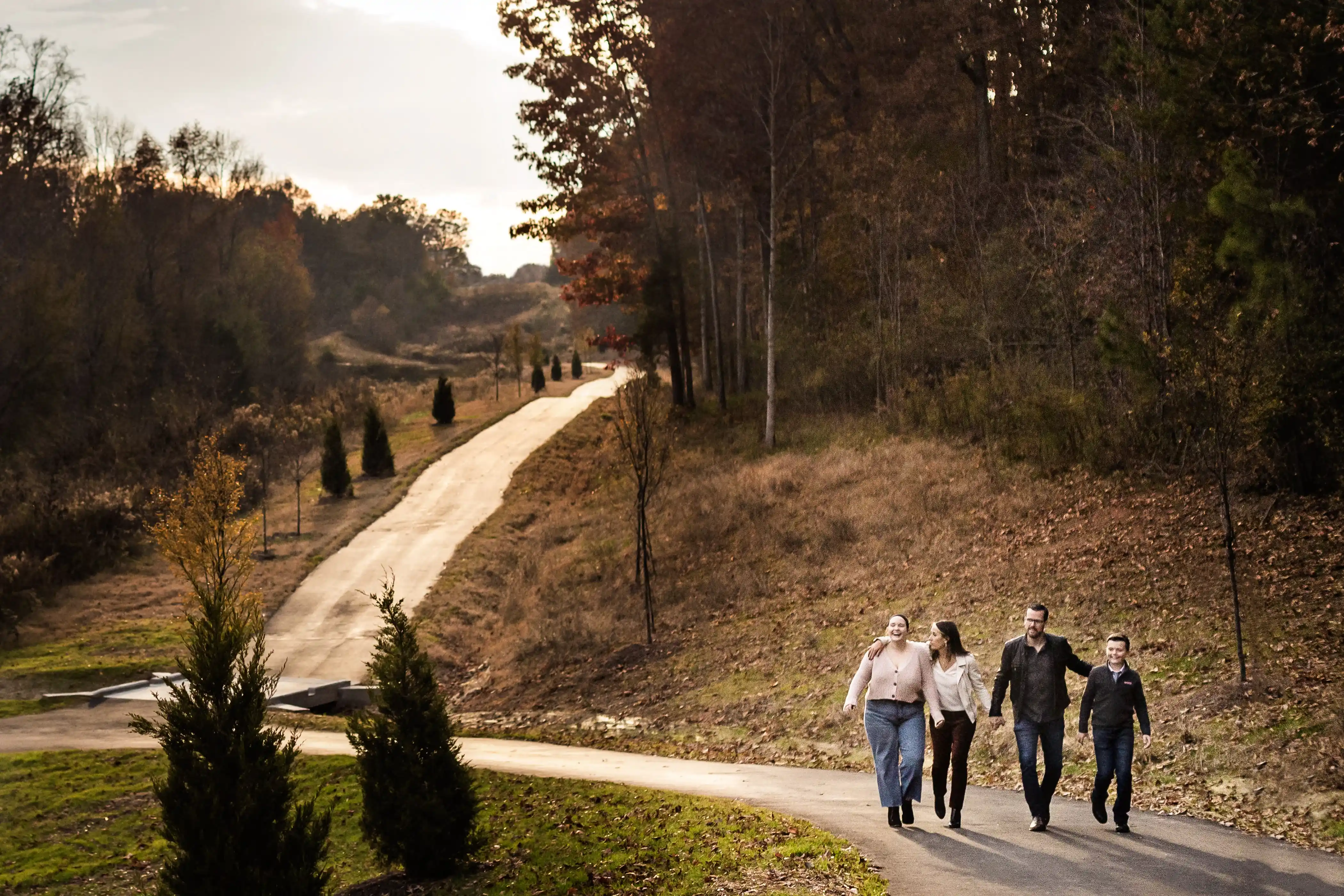 Four people walk together on a winding path through a wooded area. The path is lined with small trees, and the surrounding landscape is mostly barren, with some grass and tall trees. The sky is overcast.