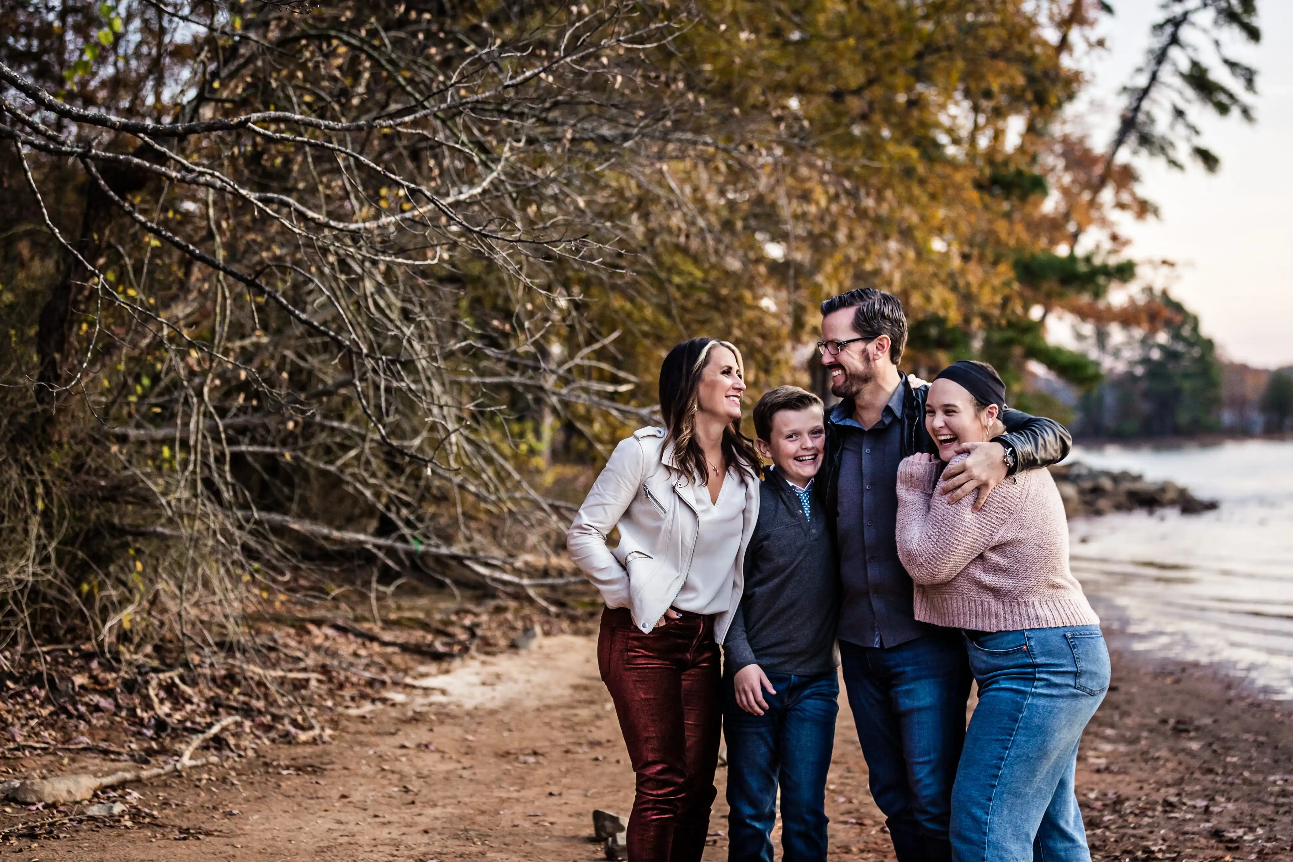 A family of four, with two adults and two children, stands on a leaf-covered path near a lake. They are smiling and embracing each other, with autumn trees in the background. The sky is clear, suggesting a mild, pleasant day.