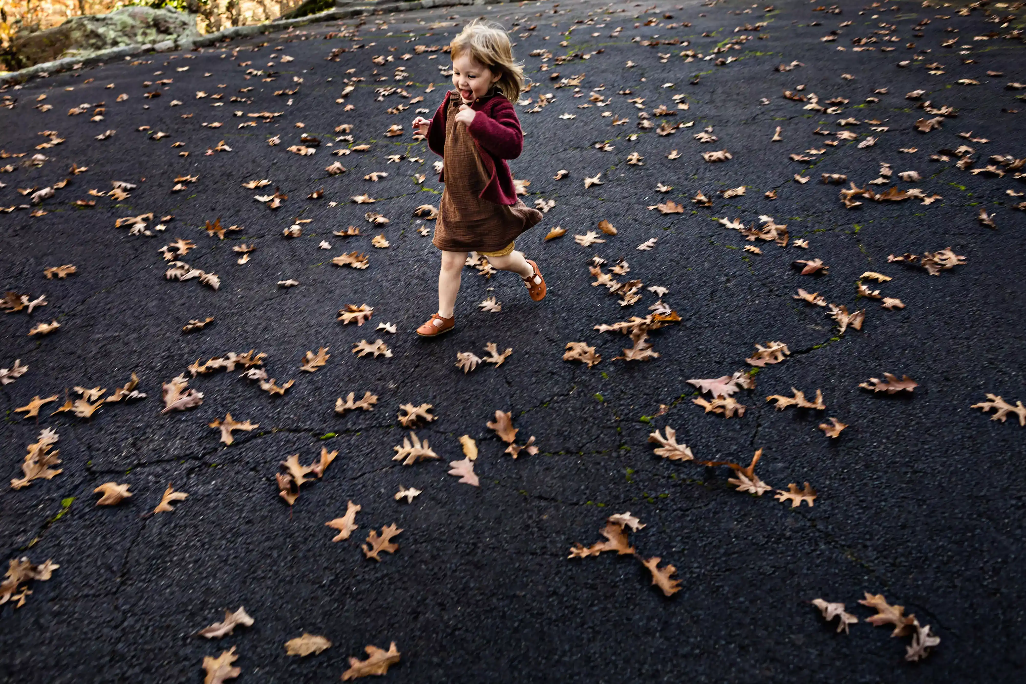 A young child in a red sweater and brown dress joyfully runs across a paved surface scattered with dry autumn leaves. The setting is outdoors and the background is blurred, emphasizing the motion and the leaves.