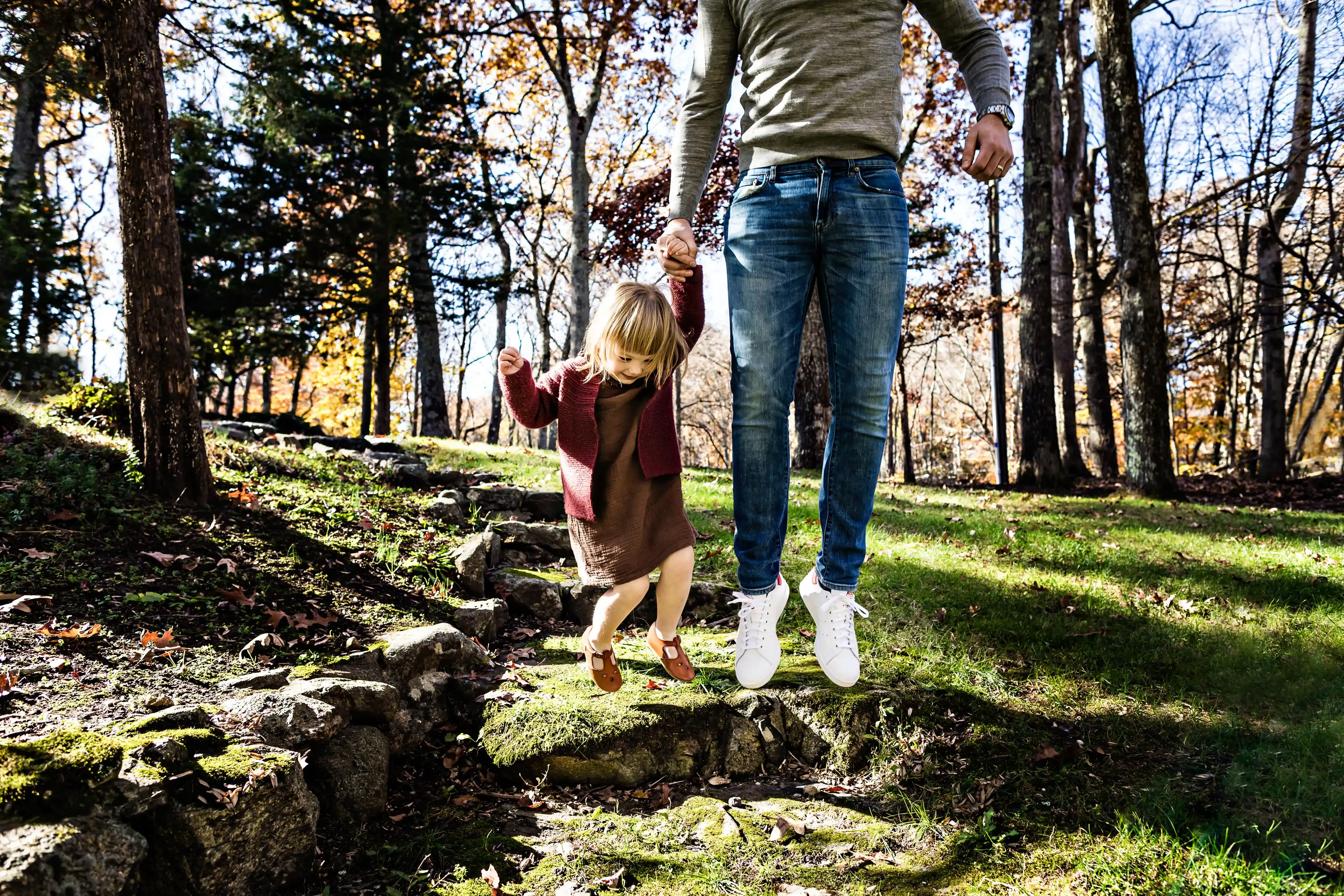 A young child and an adult holding hands joyfully jump off a small stone step in a sunlit, wooded area. The adult is wearing jeans and a green sweater, while the child is in a red coat and brown dress, with autumn trees in the background.