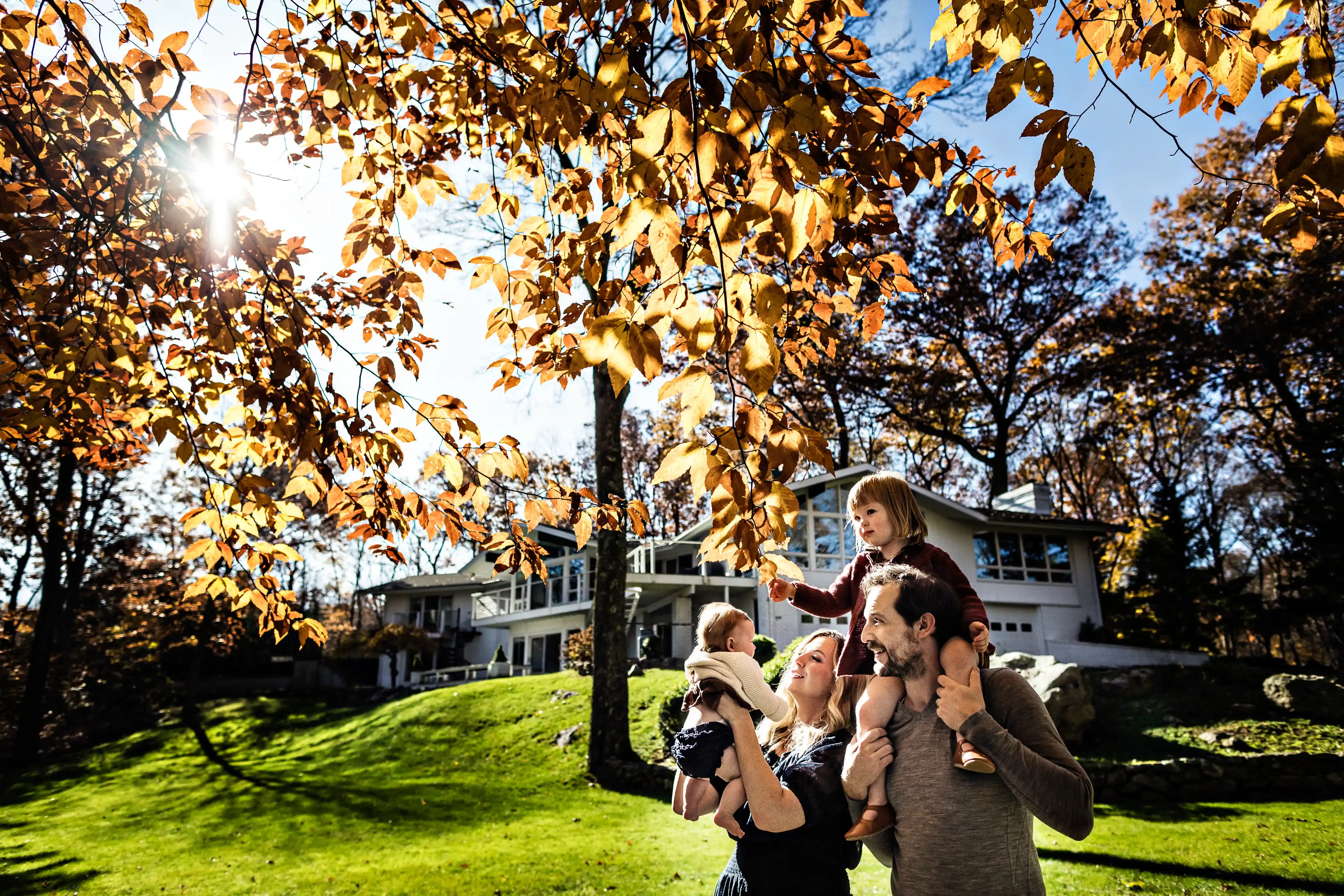A family stands in a sunlit yard surrounded by autumn trees. A woman holds a baby, while a man carries a young child on his shoulders. A modern house and a bright sky are in the background.