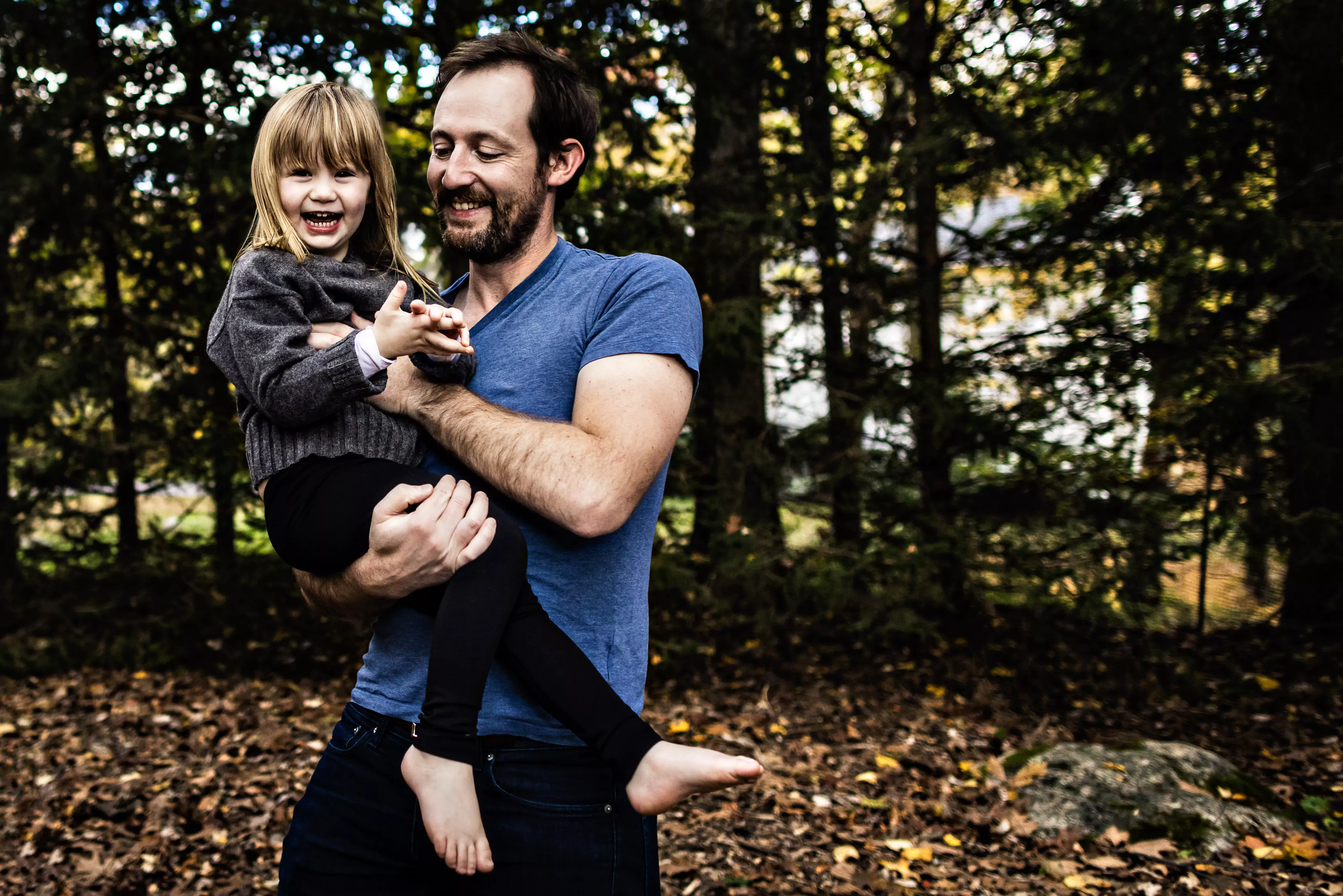 A man in a blue t-shirt joyfully holds a laughing young girl in his arms. They both have light hair. The background features trees with autumn leaves, creating a warm, natural setting.