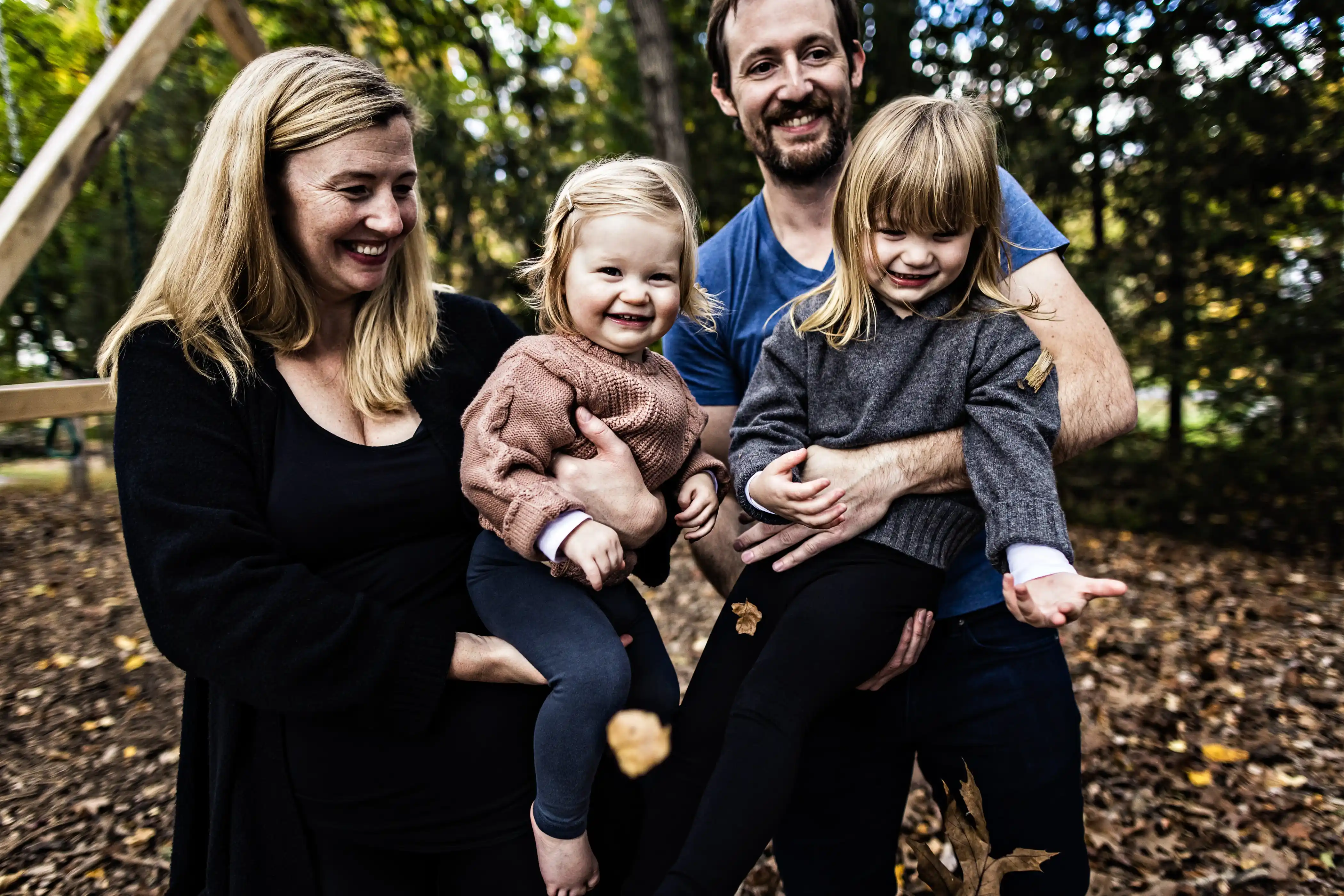 A family of four is outdoors in a wooded area, smiling and playing with fallen leaves. The adults are holding two young children, both wearing sweaters. The background is filled with trees and autumn foliage.