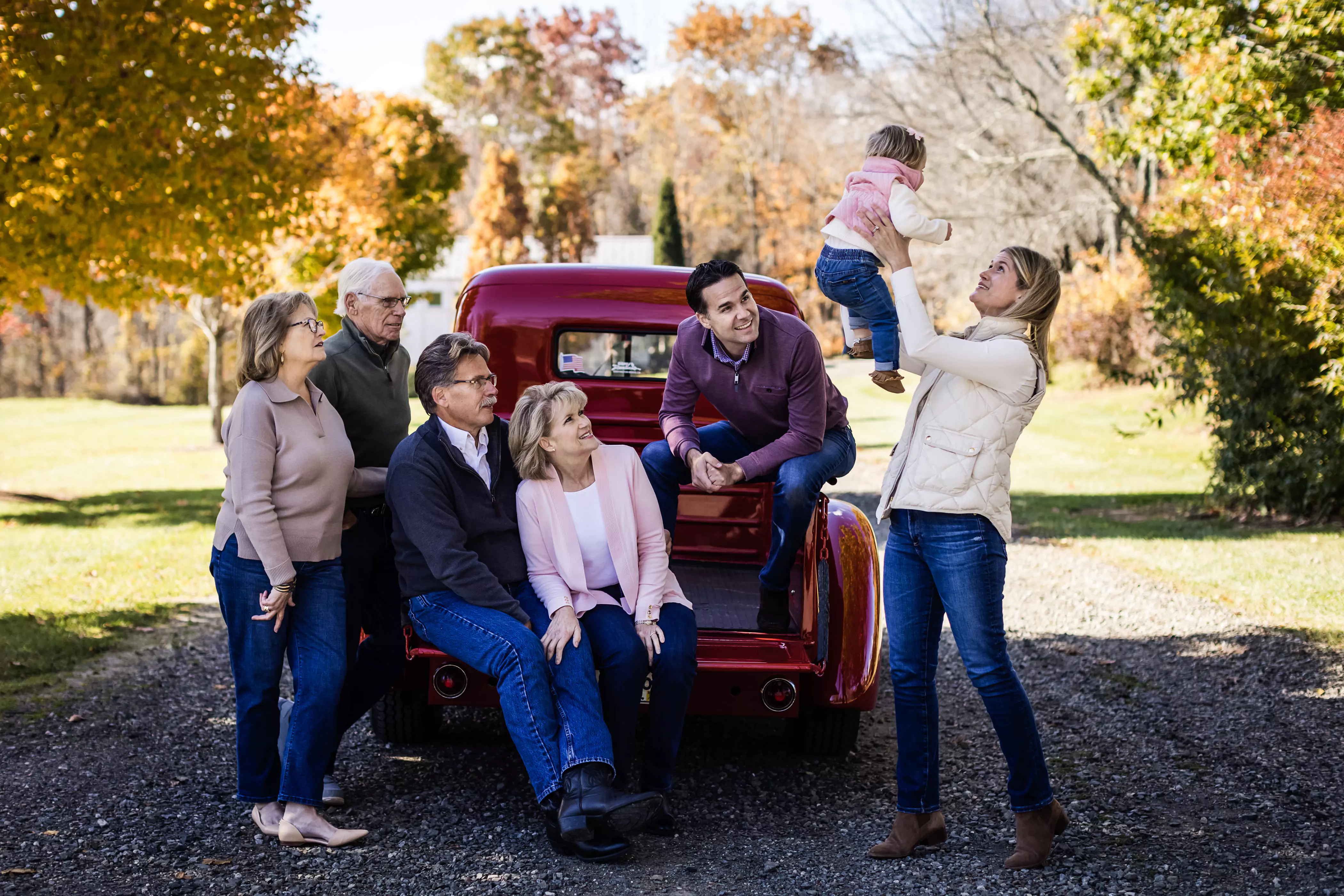 A family gathers in a park with autumn foliage, posing around a red vintage truck. Three adults sit on the truck while two stand, with a woman lifting a toddler playfully. They all wear casual clothes, enjoying a sunny, relaxed day.