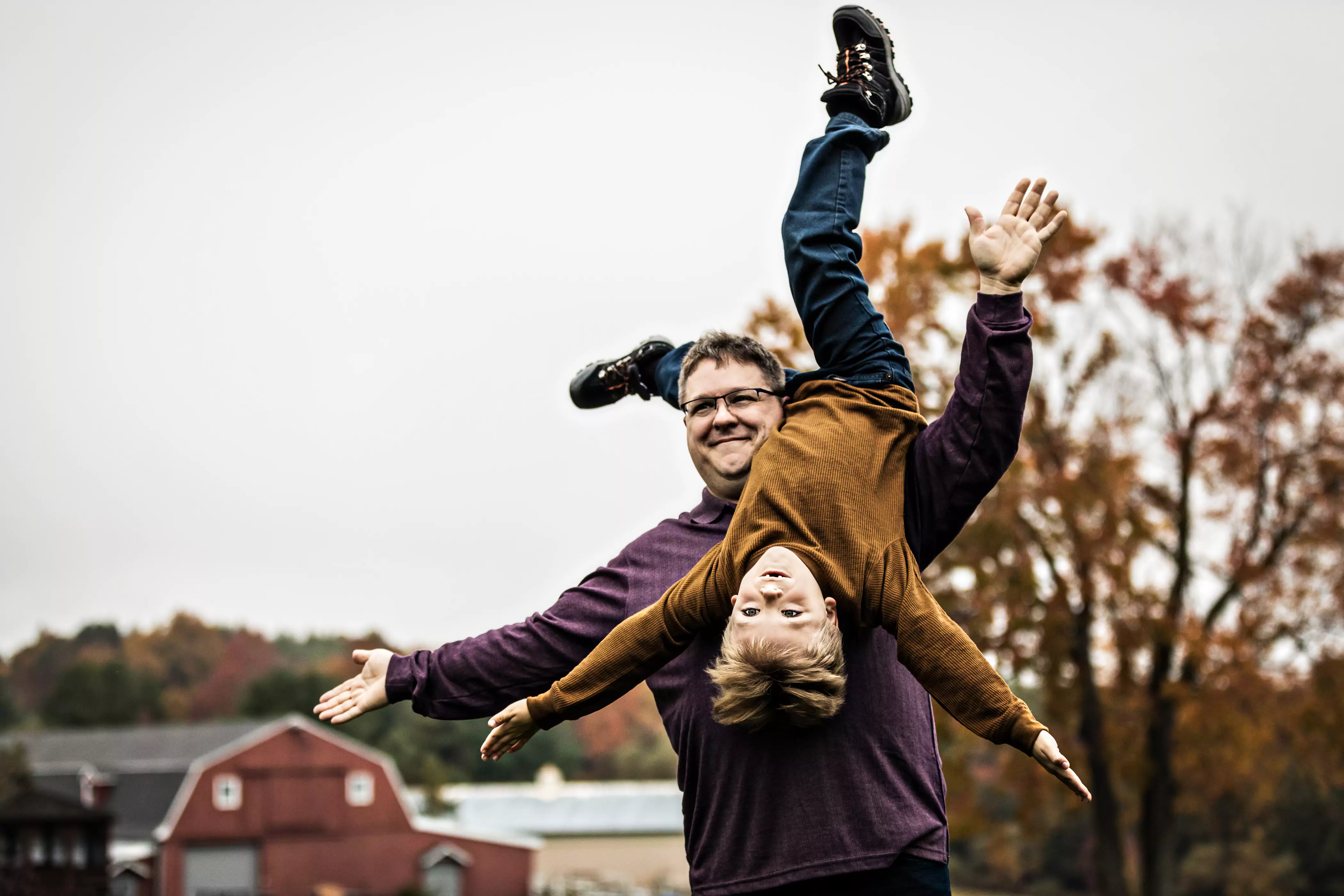 A man playfully holds an upside-down smiling child outdoors. Both have arms outstretched like an airplane. The background features autumn trees and a few red barns under a cloudy sky.