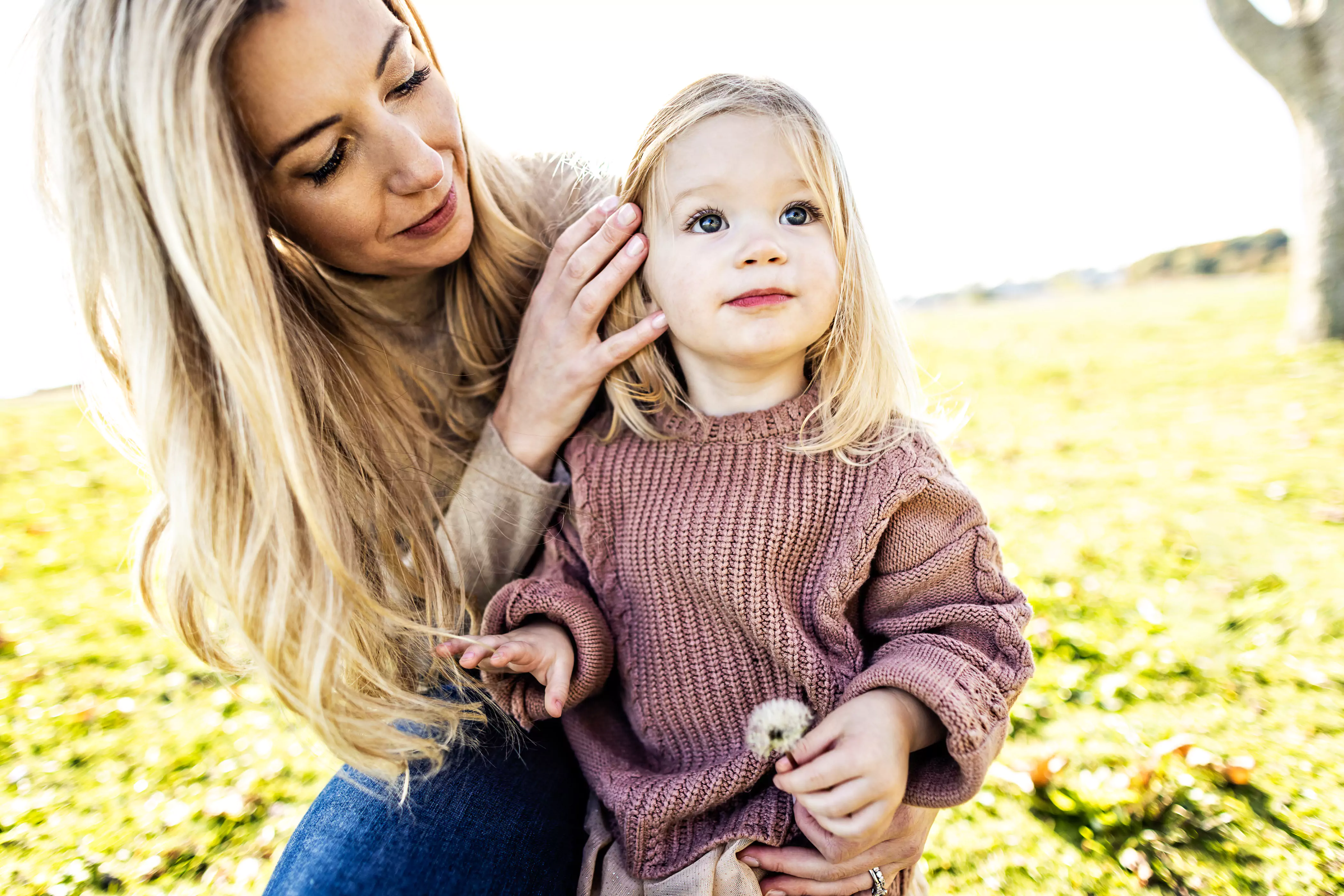 A woman with long blonde hair gently touches the hair of a small child, who is also blonde and holding a dandelion. They are outside on a sunny day, sitting on grass with blurred nature in the background.